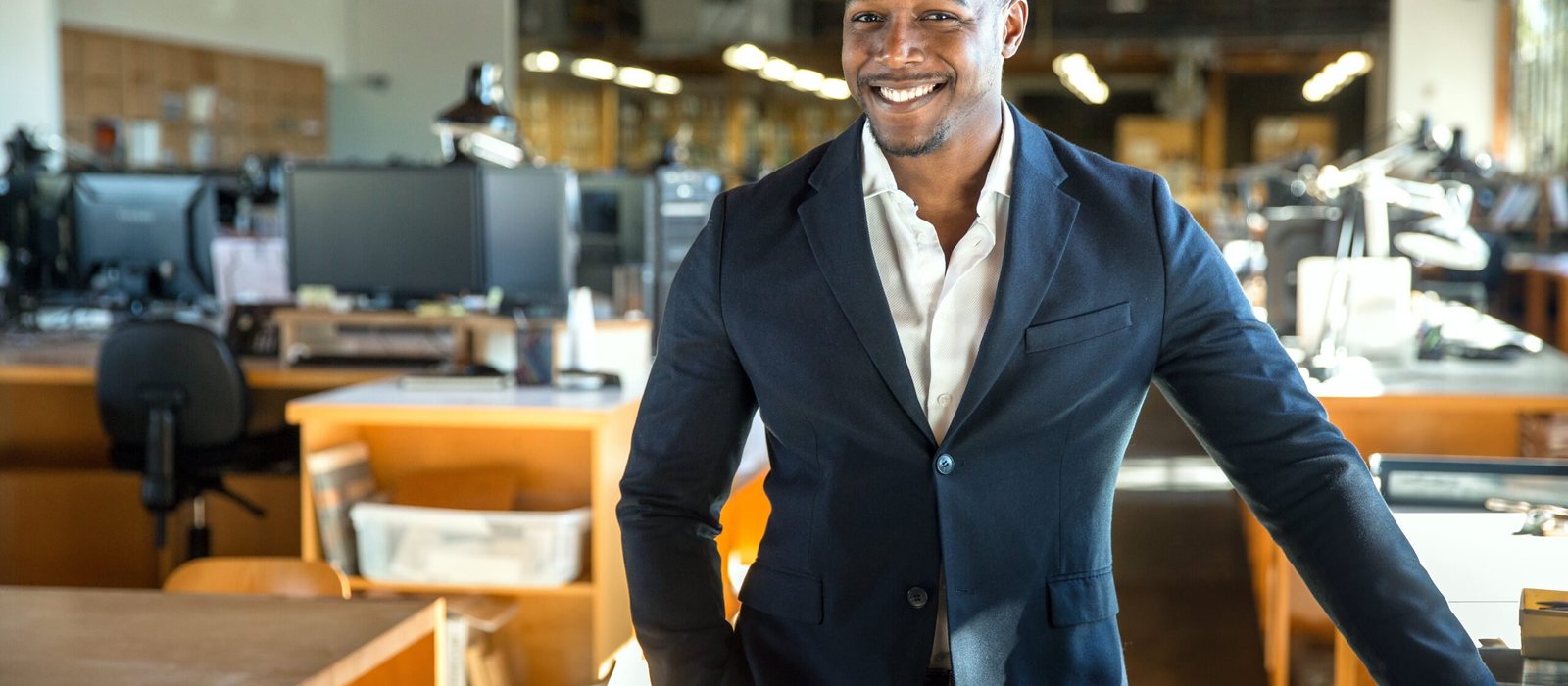 A businessman in a dark suit smiling in a bright open-plan office.