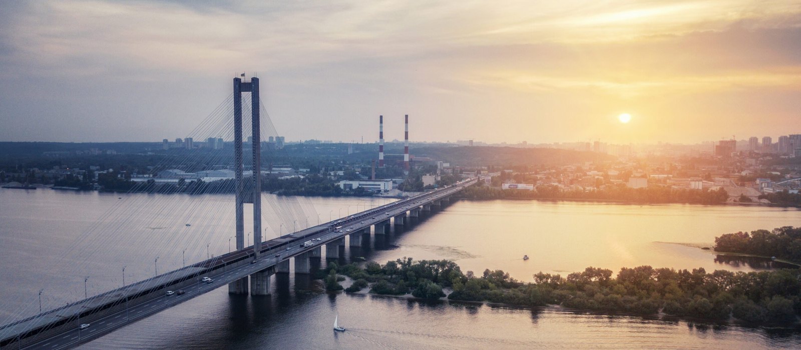 Aerial view of a cable-stayed bridge carrying cars across a wide river at sunset, with a city skyline and industrial chimneys in the background and a small sailboat on the water.
