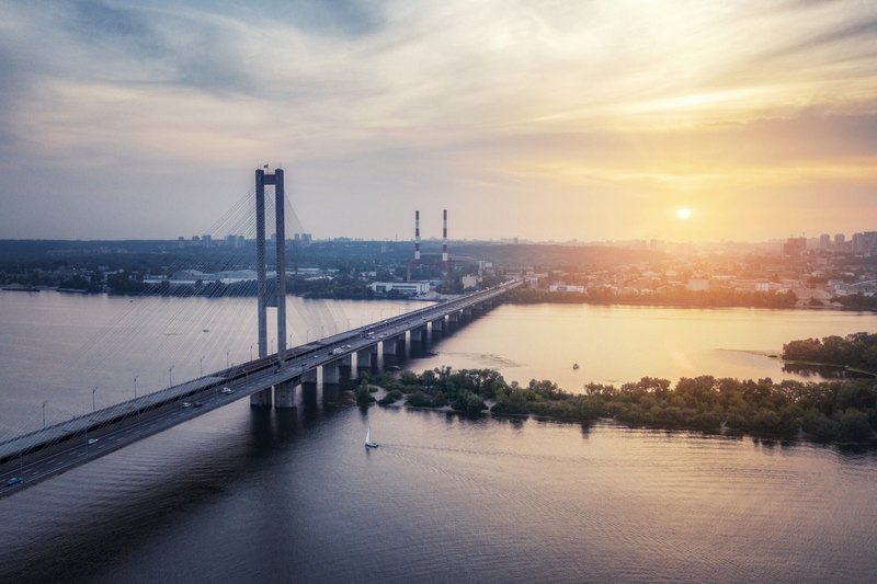 Aerial view of a cable-stayed bridge carrying cars across a wide river at sunset, with a city skyline and industrial chimneys in the background and a small sailboat on the water.