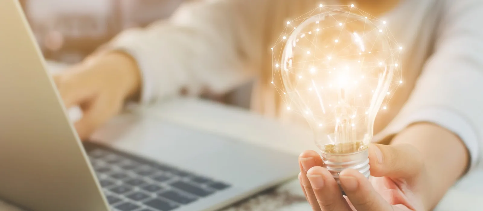 A person at a laptop holds a glowing light bulb in one open palm, with a network of dots and lines overlaid on the bulb.