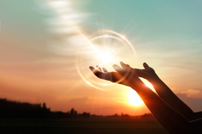 Two silhouetted hands reach upward toward a glowing light with circular lens flare against an orange and blue sunset sky above a dark horizon.