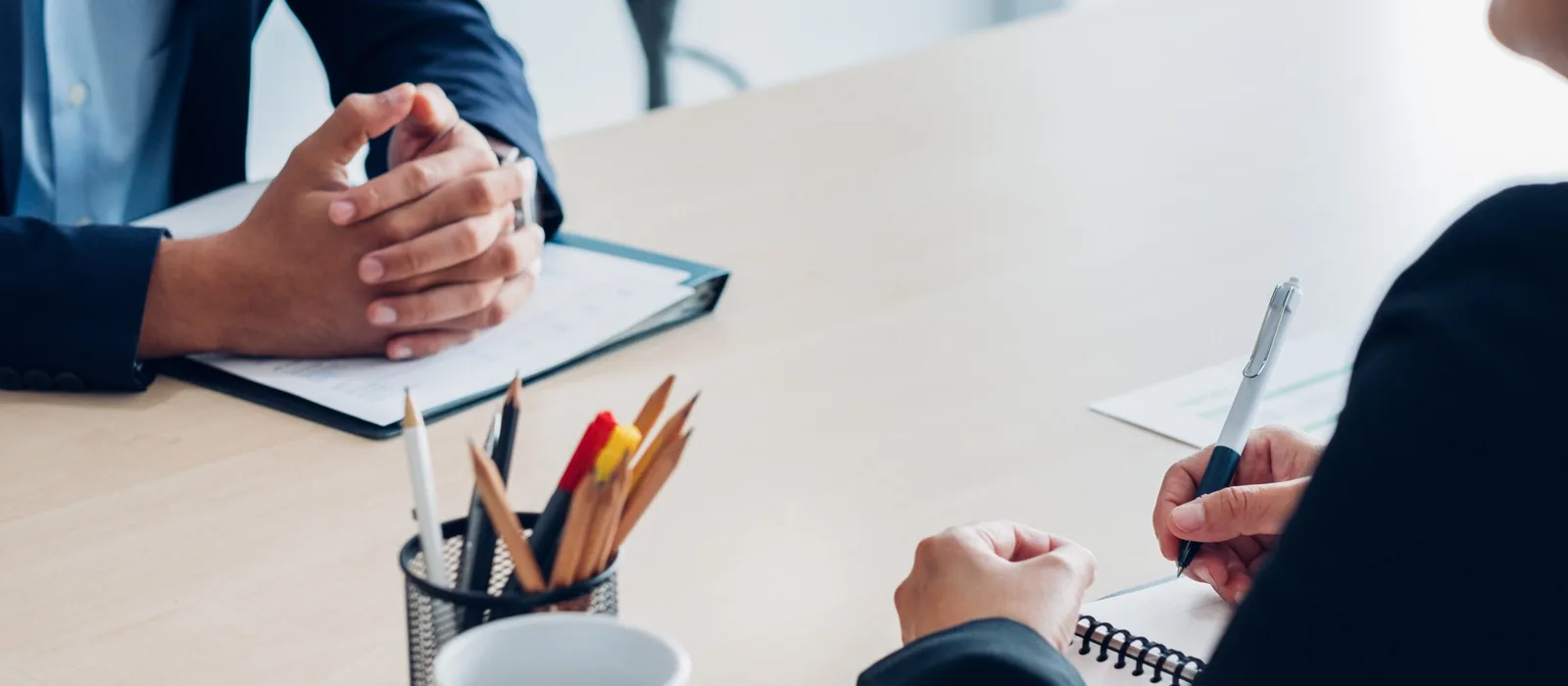 Two people in dark suits sit across a wooden table, one with clasped hands over a clipboard and the other holding a pen above a spiral notebook, beside a pencil holder and a white mug.