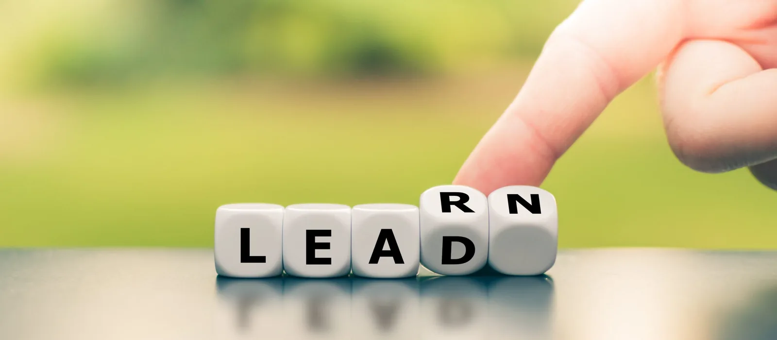 A finger turns white lettered dice on a reflective surface to change the word "LEAD" into "LEARN" against a blurred green background.
