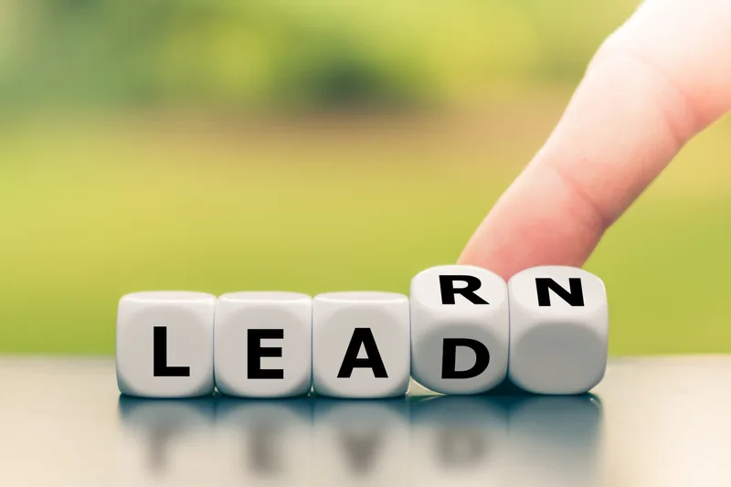 A finger turns white lettered dice on a reflective surface to change the word "LEAD" into "LEARN" against a blurred green background.
