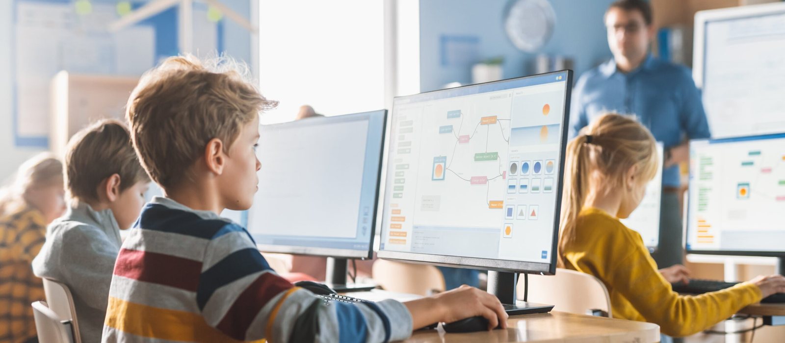 Schoolchildren working at desktop computers in a bright classroom while a teacher looks on.
