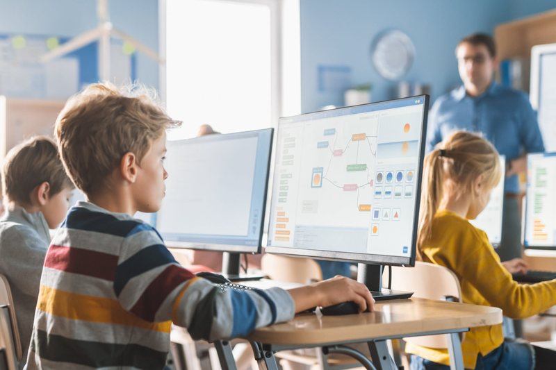 Schoolchildren working at desktop computers in a bright classroom while a teacher looks on.