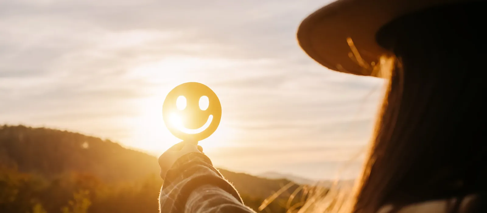 A person in a wide-brimmed hat holds up a smiley face cutout against the sun setting over wooded hills.