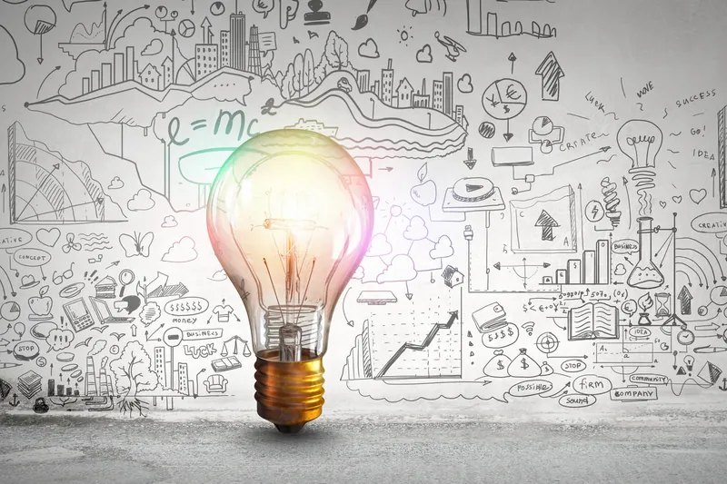 A glowing light bulb in front of a chalkboard wall covered with hand-drawn business and science diagrams.