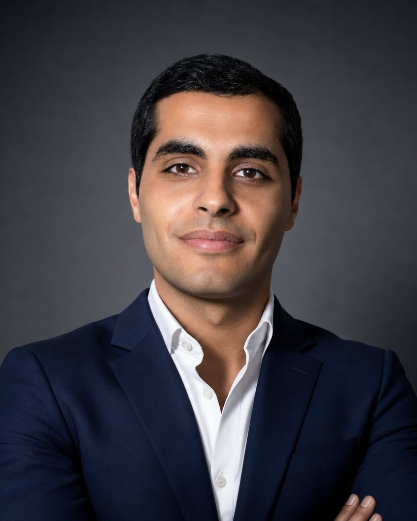 Studio portrait of Mark Elbadramany in a navy suit and white shirt against a dark grey background, arms folded, looking directly at the camera.